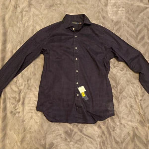 Ralph Lauren Skjorta - En mörkblå prickig skjorta från Ralph Lauren. Skjortan är helt ny och aldrig använd. Tags sitter kvar och även extra knappar medföljer som går att se på bild 4. Den fungerar bra att ha på sig under en kostym eller bara som den är.