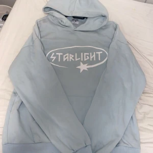 Ljusblå hoodie från Starlight - Säljer en ljusblå hoodie från Starlight med vit tryckt logga och stjärndetalj på bröstet. Tröjan har huva, långa ärmar och en klassisk känguruficka framtill. Perfekt för dig som gillar en chill och stilren look.