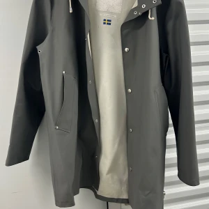 Grå regnjacka med huva och snörning - Snygg grå regnjacka med huva och vita snören. Jackan har tryckknappar framtill och två stora fickor med lock. Perfekt för regniga dagar och enkel att matcha med olika outfits. Klassisk och stilren design.