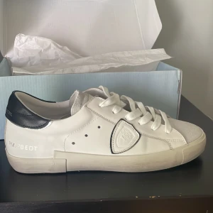 Vita sneakers från Philippe Model - Säljer ett par snygga vita Philippe Model sneakers med svarta detaljer på hälen och vit snörning. Skorna består av både mocka och skinn. Skorna är ALDRIG ANVÄNDA utan bara provade! Säljer pga att de inte passade i storlek. Ny pris 3300 kr mitt pris 2500. Priset inte hugget i sten! Hör gärna av dig vid eventuella frågor!