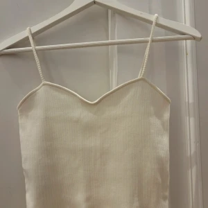 Vit ribbad linnetopp med smala band - Supersöt vit ribbad linnetopp med smala axelband och en lätt hjärtformad urringning. Toppen är croppad och har en enkel, stilren look som passar perfekt till jeans eller kjol. Mjuk och stretchig för en bekväm passform.