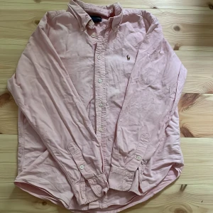Ljusrosa skjorta från Ralph Lauren - Klassisk ljusrosa skjorta från Ralph Lauren med button-down krage och broderad logga på bröstet. Skjortan har långa ärmar, knappar framtill och är tillverkad i mjuk bomull. Perfekt för en clean och stilren look.