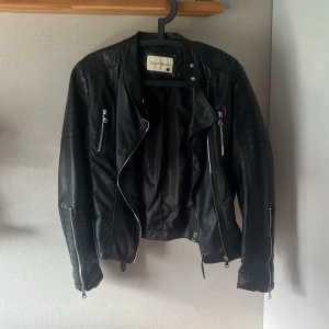 Svart skinnjacka från King of Fashion - Snygg svart skinnjacka med coola detaljer som dragkedjor på ärmar och bröst, samt quiltade partier på axlarna. Jackan har klassisk bikerstil med sned dragkedja och flera fickor. Perfekt för dig som vill ha en edgy look. Storleken är XL men passa mig o min kompis som har S/M, den har ett hål inne i jackan i de tunna lagret men inget som syns när den är på.