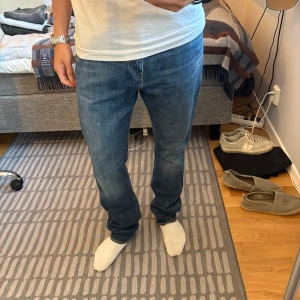 Levis 501 blå jeans straight fit - Vintage Levis 501 jeans i blå denim med raka ben och normal passform. Jeansen har fem fickor, knappgylf och tydliga Levis-detaljer som röd etikett och patch bak. Perfekta för en avslappnad och tidlös look. På byxorna står det L34W34, men personligen tycker jag längden passar för någon med storlek L32