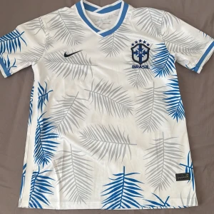 Vit Brasilien fotbollströja Nike S - Snygg vit Brasilien fotbollströja från Nike med blå och grå palmbladsmönster. Tröjan har korta ärmar, blå detaljer vid krage och ärmslut samt broderat CBF-märke på bröstet. Tillverkad i lätt och ventilerande Dri-FIT-material.