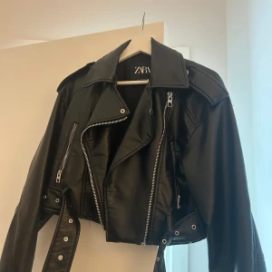 Svart skinnjacka från Zara - Snygg svart skinnjacka från Zara med klassisk bikerstil. Jackan har silvriga dragkedjor, bred krage och bälte i midjan. Croppad modell med långa ärmar och coola detaljer.