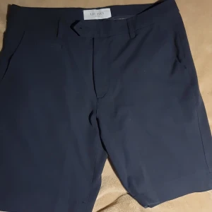 Mörkblå shorts från Les Deux - Stilrena mörkblå shorts från Les Deux, modell Como Light. Klassisk design med fickor fram och bak, bälteshällor och knappstängning. Sköna och lätta i polyesterblandning, perfekta för varma dagar. Snygg passform som funkar till både t-shirt och skjorta.