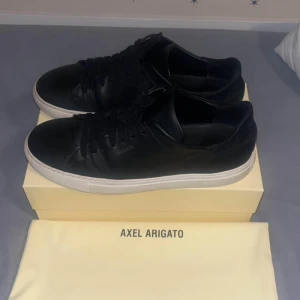  Axel Arigato skor - Stilrena svarta sneakers från Axel Arigato i skinn med vit platt sula. Klassisk låg modell med rund tå och svarta snören. Perfekta för dig som gillar minimalistisk och clean design. Kommer med originalkartong och dustbag.