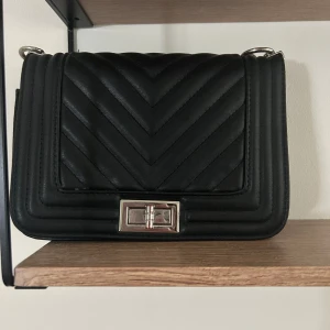 Svart quiltad axelväska med kedja - Snygg svart axelväska med quiltad design och silvrig kedjerem. Väskan har ett stilrent spänne framtill och en kompakt, boxig form som ger en modern vibe. Perfekt för dig som vill ha en trendig accessoar med plats för det viktigaste.