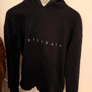 Svart hoodie Jack & Jones Originals - Svart hoodie från Jack & Jones Originals med vit text över bröstet. Klassisk känguruficka framtill, huva med dragsko och mjukt material på insidan. Perfekt för en chill och avslappnad stil.