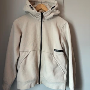 Beige hoodie från Sail Racing - Snygg beige hoodie från Sail Racing med dragkedja framtill och två stora fickor. Jackan har huva med snörning och är fodrad med mjuk fleece på insidan. Perfekt för chill dagar och enkel att matcha med det mesta. Hör gärna av er vid frågor!