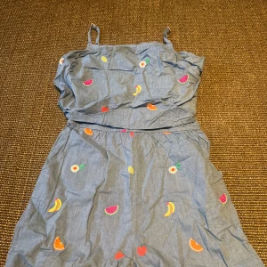 Blå byxdress med fruktbroderi - Söt blå byxdress med tunna axelband och broderade frukter som bananer, apelsiner, jordgubbar och vattenmelon. 