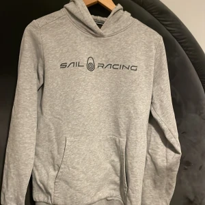 Grå hoodie från Sail Racing - Säljer en ljusgrå hoodie från Sail Racing med svart logga tryckt på bröstet. Tröjan har huva och känguruficka framtill. Perfekt för en avslappnad stil och enkel att matcha med det mesta.