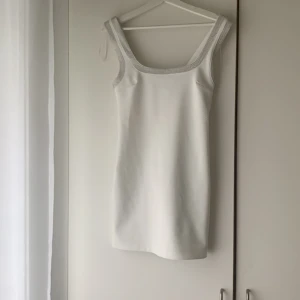 Vit kort klänning med strassdetaljer från Zara - Säljer en vit kort klänning från Zara i storlek XS. Klänningen är ärmlös och har snygga strassdetaljer längs ringningen och axelbanden. Perfekt för dig som vill sticka ut med en stilren och glittrig look. Jag har haft den på mig en gång så den är helt ny.