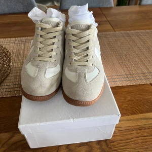 Beige och vita sneakers från Maison Margiela - Säljer ett par stilrena sneakers från Maison Margiela i beige mocka och vitt skinn med gummisula. Klassisk design med rund tå och platt sula, perfekta för en clean och trendig look.