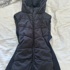 Svart quiltad dunväst med huva från Zara - Säljer en svart quiltad dunväst från Zara med huva och dragkedja framtill. Västen har en figurnära passform och är perfekt för lager-på-lager-stil. Snygg och enkel att matcha till olika outfits. 