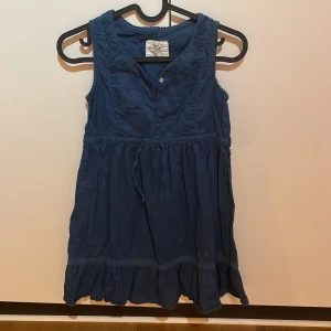 Marinblå ärmlös klänning från L.O.G.G. - Söt marinblå ärmlös klänning från L.O.G.G. med knappar framtill och dekorativa broderier upptill. Klänningen har volang nertill och ett knytband i midjan för en fin passform. Perfekt för varma dagar och enkel att styla.