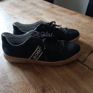Svarta sneakers från Björn Borg - Svarta sneakers från Björn Borg med vit sula och vita detaljer. Skorna har snörning och en tydlig logga på sidan. Ovandelen är i textil och insidan är ljus. Perfekta för en avslappnad stil. Dom är i jätte bra skick lite smusiga kanske men det ända problemey är att skosulorna är trasiga man man kan säker köpa nya för en billig peng.