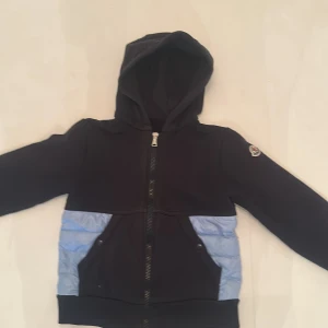Svart och ljusblå hoodiejacka från Moncler - Snygg hoodiejacka från Moncler i svart med ljusblå quiltade partier på nedre delen och baksidan. Jackan har huva, dragkedja framtill och två fickor med knapp. Moncler-logga på ärmen. Perfekt för dig som vill ha en sportig och stilren look.