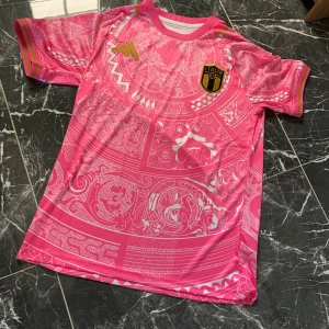 Italien-inspirerad rosa fotbollströja - Säljer en rosa fotbollströja med vita mönster och guldiga detaljer. Tröjan har korta ärmar och ett tryckt Italien-emblem på bröstet. Unik design med dekorativa mönster och inslag av guld på ärmarna.