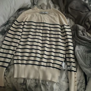 Randig tröja från ZARA med knappar - Säljer en vit långärmad tröja från ZARA med marinblå ränder och dekorativa guldfärgade knappar vid ärmsluten. Tröjan har en klassisk passform och är perfekt för en stilren look.