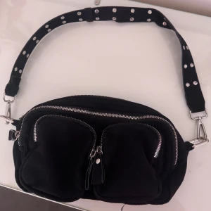 Svart axelväska från Gina Tricot - Säljer en svart axelväska från Gina Tricot med bred axelrem dekorerad med silvriga nitar. Väskan har två stora ytterfickor med dragkedja och en innerficka. Perfekt för att bära med sig det viktigaste.