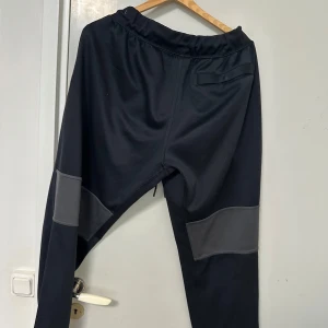 Svarta mjukisbyxor från Nike med gråa detaljer - Svarta mjukisbyxor från Nike med gråa paneler på knäna och vit Nike-logga på vänster lår. Byxorna har elastisk midja med dragsko och en bakficka. Perfekta för träning eller chill.