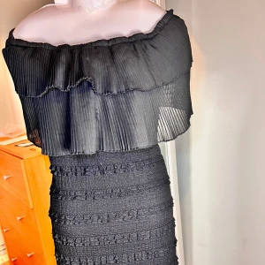 Svart offshoulder långklänning vågigt  - Säljer en svart långklänning med offshoulder-modell och plisserad volang upptill. Klänningen har en figurnära passform och struktur i tyget som ger en unik look. Perfekt för dig som vill sticka ut med en elegant och trendig stil.