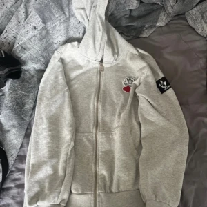 Grå hoodie från Moncler Grenoble - Säljer en ljusgrå hoodie från Moncler Grenoble med dragkedja framtill. Tröjan har huva, två fickor och broderade Moncler-loggor på bröstet och ärmen. Perfekt för en avslappnad och stilren look.