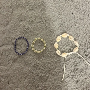 Tre armband med pärlor och snäckor - Säljer tre armband: ett med blå och vita pärlor, ett med små vita blommor och gröna pärlor, samt ett med vita snäckor och justerbart band. Perfekta accessoarer för en somrig look!