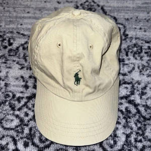Beige keps från Polo Ralph Lauren - Klassisk beige keps från Polo Ralph Lauren med mörkgrönt broderat logotyp framtill och 'POLO' broderat baktill. Justerbar rem baktill för perfekt passform. Tillverkad i bomull och passar till många olika stilar.