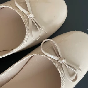 Beige ballerinaskor med rosett från Nelly - Säljer ett par stilrena beige ballerinaskor från Nelly med söt rosettdetalj framtill. Skorna har rund tå och platt sula, perfekta för en enkel och klassisk look. Materialet är syntet och färgen är ljusbeige. Nyskick - aldrig använda😇