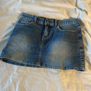 Lågmidjad jeanskjol från Only Jeans - Säljer en lågmidjad blå jeanskjol från Only Jeans med femficksmodell och synliga sömmar. Kjolen har bälteshällor, knapp- och dragkedjestängning samt bakfickor med dekorativa sömmar. Perfekt för dig som gillar en tidlös denimlook.