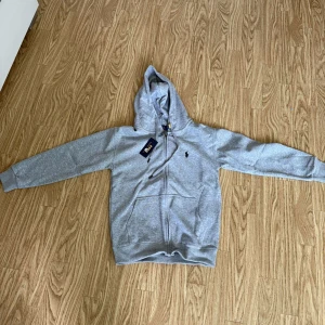 Grå kofta från Polo Ralph Lauren - Säljer en klassisk grå kofta från Polo Ralph Lauren med dragkedja framtill och huva. Tröjan har långa ärmar, två fickor och den ikoniska lilla broderade loggan på bröstet. Perfekt för en avslappnad stil.