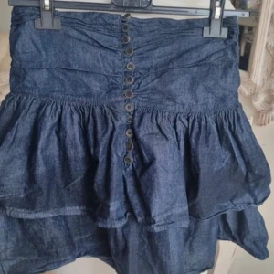 Jeanskjol - Supersöt mörkblå jeanskjol från Zara Woman med volanger och knappar på sidan. Kjolen är jätte fin med en tex vit T-shirt till och passar perfekt nu till sommaren en vardag eller utekväll. Storlek är m men passar mig som brukar ha 34-36 i storlek! Inga defekter.