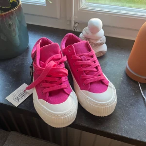 Rosa sneakers från Juicy couture - Säljer ett par snygga rosa sneakers från Juicy couture med vit platt sula och rund tå. Skorna har snörning och är i textilmaterial, perfekta för att liva upp din outfit. Skorna är helt oanvända och har prislappen kvar! 