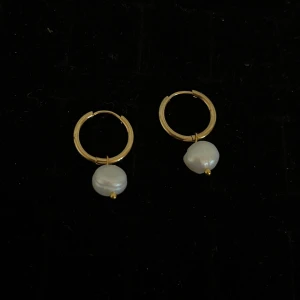 Guldiga örhängen med pärlor - Säljer ett par stilrena guldiga örhängen med vita pärlor. Perfekta för att ge en elegant touch till din look. Runda ringar med hängande pärldetalj.