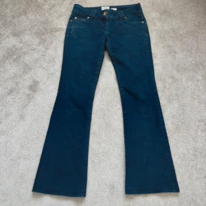 Blå bootcut jeans med låg midja - Säljer ett par klassiska blå bootcut jeans med låg midja. Jeansen har fem fickor, gylf med dragkedja och knapp samt snygga detaljer i guld vid fickorna. Perfekta för dig som gillar en retroinspirerad look.