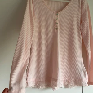 Ljusrosa ribbad topp med spets från Janina - Supersöt ljusrosa långärmad topp från Janina med ribbad struktur, spetsdetaljer vid halsringning och nederkant samt små knappar framtill. Perfekt för en romantisk look! 💗