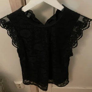 Svart spetsblus från Pieces - Säljer en svart blus från Pieces i storlek XS. Blusen är ärmlös med vackra spetsdetaljer och blommigt mönster. Perfekt för dig som gillar feminina och stilrena plagg med extra detaljer vid ärmslut och hals.