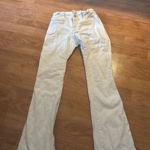 Vita bootcut-byxor från H&M - Säljer ett par vita bootcut-byxor från H&M med låg midja och justerbar midja. Byxorna har klassisk femficksdesign och stängs med dragkedja och knapp. Perfekta för dig som gillar en lite vidare passform nertill.
