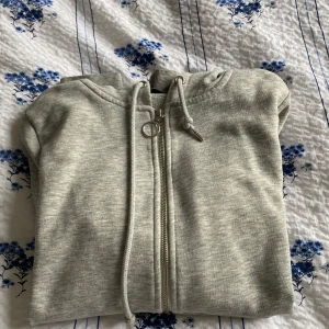 Ljusgrå hoodie med dragkedja - Ljusgrå hoodie med dragkedja och huva. Tröjan har snörning i huvan och ribbade muddar vid ärmslut och nederkant. Perfekt för en avslappnad stil och enkel att matcha med andra plagg.