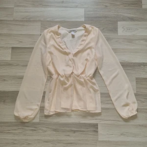 Beige blus från NLY Trend - Säljer en ljusbeige blus från NLY Trend i storlek 36. Blusen har långa ärmar, markerad midja och v-ringad omlottdesign. Perfekt för en stilren och feminin look.