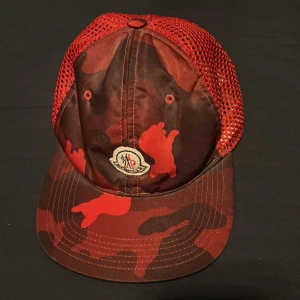 Röd camo keps från Moncler - Säljer en röd och mörkröd keps från Moncler med camouflagemönster och mesh baktill. Justerbart spänne i metall med Moncler-logga. Perfekt för dig som vill sticka ut med en snygg accessoar. out off stock överallt, använd 3-4 gånger. 