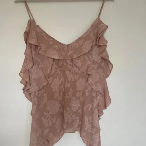 Rosa volanglinnetopp från Zara - Säljer en ljusrosa linnetopp från Zara med tunna axelband och volangdetaljer längs fram- och sidorna. Toppen har ett subtilt blommönster och är perfekt för varma dagar.