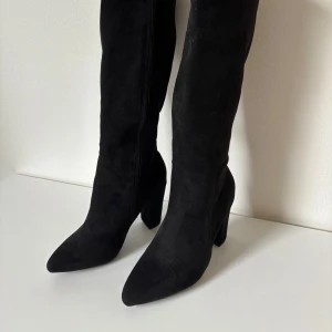 Svarta höga stövlar med klack - Säljer ett par svarta höga stövlar i mocka-imitation med dragkedja på insidan (silver) och ca 10 cm klack. Endast provade. Skorna är storlek 40 men passar som 38/39.