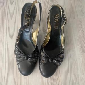 Novita klackskor - Snygga svarta pumps från Novita med öppen häl och justerbart spänne. Skorna har dekorativa nitar och en rosett framtill samt guldfärgad insida. Perfekta för dig som vill ha en elegant och unik sko!