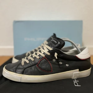Svarta sneakers från Philippe Model  - Snygga svarta sneakers från Philippe Model med vita snören och vit häl. Skorna har en unik sköld-detalj på sidan med röd kant och en platt vit sula. Perfekta för en stilren och avslappnad look. Storlek 43. Bra skick! Nypris runt 3000 kr