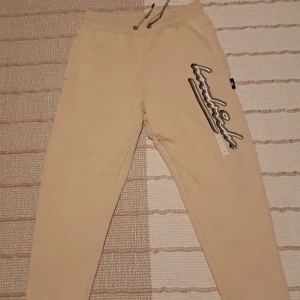 Beige mjukisbyxor med tryck och snörning - Säljer ett par beige mjukisbyxor med coolt tryck längs ena benet och snörning i midjan. Perfekta för en avslappnad stil och sköna att ha på sig hemma eller till vardags.