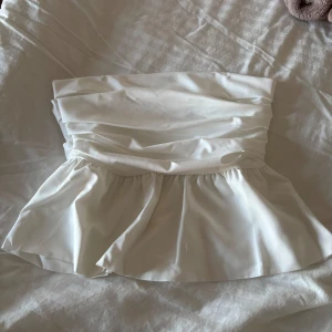 Vit volang tubtopp - Säljer en söt vit tubtopp med volang nertill. Toppen har en draperad design upptill och är axelbandslös, perfekt för sommardagar. Materialet känns mjukt och följsamt.